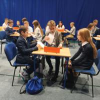 Grupy uczniów siedzą w trzyosobowych zespołach przy stolikach z nazwą szkoły