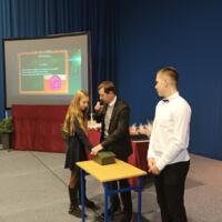 Dziewczynka ma rękę włożoną do zamkniętego pudełka. Prowadzący trzyma w ręku mikrofon i uśmiecha się do dziewczynki.