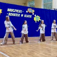 Dziewczynki biorące udział w akademii wykonują układ choreograficzny do nowoczesnej muzyki. Ubrane są w luźne stroje.