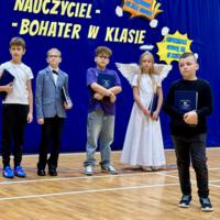 Uczniowie czytający wiersze, w tle na niebieskiej kotarze barwy narodowe oraz napis "Nauczyciel bohater w klasie"