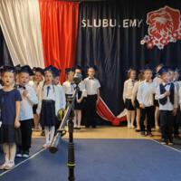 Uczniowie klas pierwszych ubrani galowo w białe koszule z granatowymi wstążkami pod szyją oraz dziewczynki granatowe lub czarne spódniczki, chłopcy granatowe lub czarne spodnie. Na głowach dzieci mają granatowe birety. W tle  na granatowej kotarze w centrum biały napis ślubujemy. Po lewej stronie barwy narodowe oraz przypięte biało czerwone kwiaty z papieru. Po prawej stronie wycięta z papieru i przymocowana do kotary głowa orła. Dzieci stoją na granatowej wykładzinie. Recytują przygotowany scenariusz.