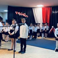 Uczniowie klas pierwszych ubrani galowo w białe koszule z granatowymi wstążkami pod szyją oraz dziewczynki granatowe lub czarne spódniczki, chłopcy granatowe lub czarne spodnie. Na głowach dzieci mają granatowe birety. W tle  na granatowej kotarze w centrum biały napis ślubujemy. Po lewej stronie barwy narodowe oraz przypięte biało czerwone kwiaty z papieru. Po prawej stronie wycięta z papieru i przymocowana do kotary głowa orła. Dzieci stoją na granatowej wykładzinie. Recytują przygotowany scenariusz.
