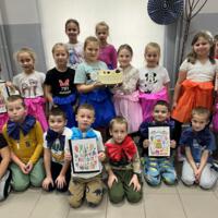 Dzieci ustawione są do wspólnego zdjęcia. W centralnej części dwie dziewczynki trzymają w ręku tort urodzinowy, kilkoro dzieci ma w rękach własnoręcznie wykonane kartki urodzinowe. 